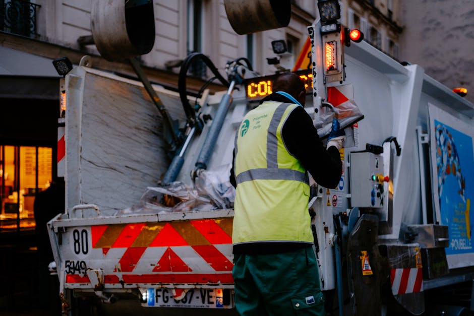 A worker wearing a high-visibility yellow safety vest and dark clothing operates a mechanical waste collection vehicle on an urban street during dusk or early evening. The vehicle's rear compartment i
