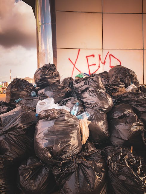 A worker wearing a high-visibility yellow safety vest and dark clothing operates a mechanical waste collection vehicle on an urban street during dusk or early evening. The vehicle's rear compartment i