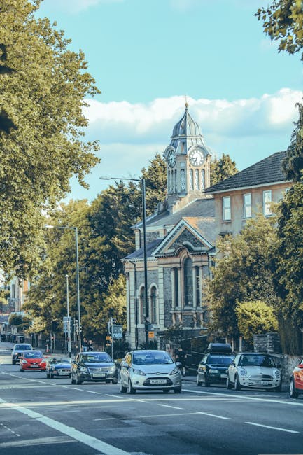 A street scene in Wandsworth featuring a church with a tall clock tower and spire in the background, surrounded by mature trees with green foliage. Several parked cars line the pavement in front of th