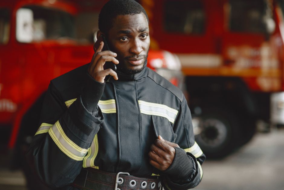 A male firefighter with dark skin, short black hair, and a serious expression, is holding a mobile phone to his ear with his right hand and adjusting his black protective uniform with his left hand. T
