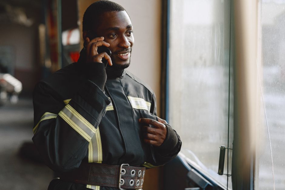 A male firefighter with dark skin, short black hair, and a serious expression, is holding a mobile phone to his ear with his right hand and adjusting his black protective uniform with his left hand. T